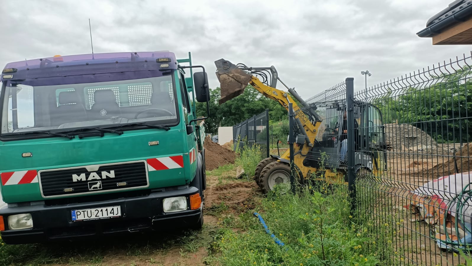 Żółta miniładowarka wykonuje prace ziemne obok zielonej ciężarówki MAN na tle ogrodzenia panelowego i sterty ziemi. Widoczne elementy instalacji.