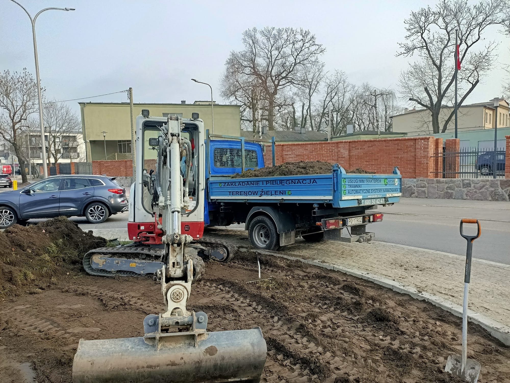 Mini koparka i wywrotka z ziemią podczas prac ziemnych. Widoczna łopata i ślady gąsienic. Na wywrotce napis: Zakładanie i pielęgnacja terenów zieleni.