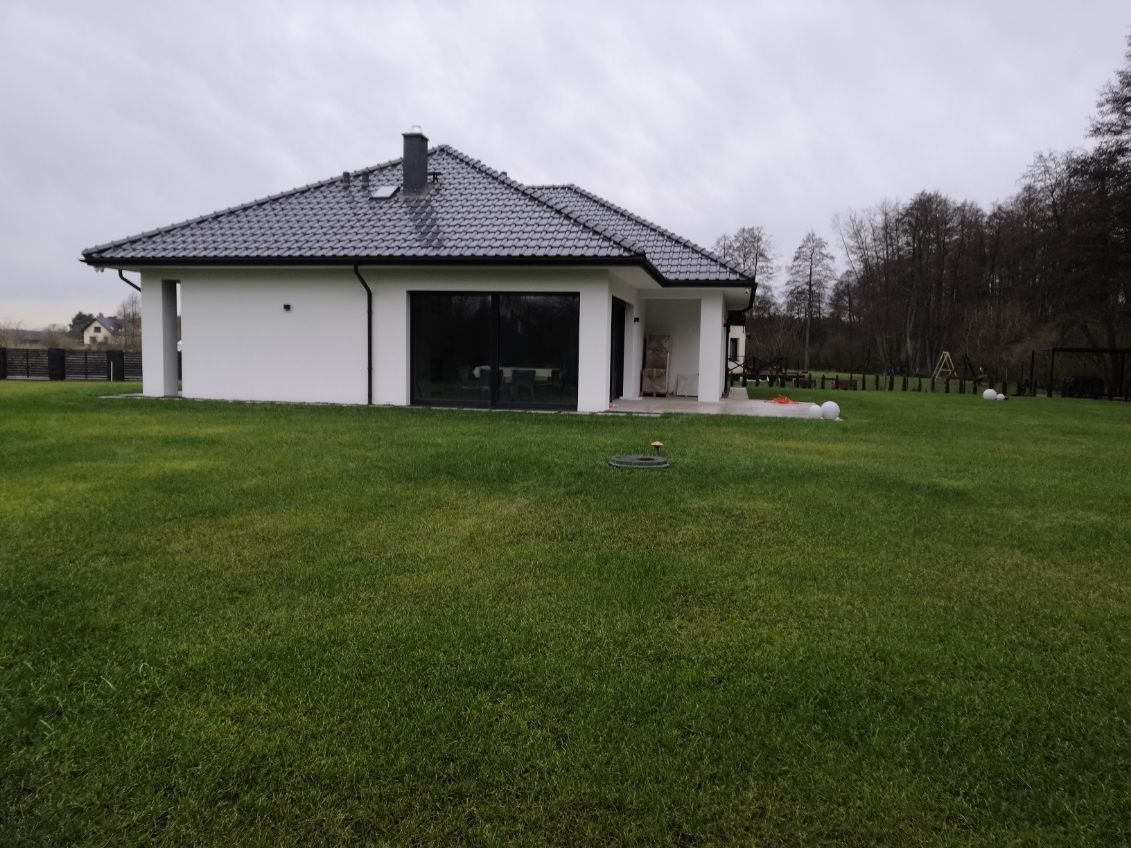 Parterowy dom z białymi ścianami i grafitowym dachem z dachówki ceramicznej, duże okno przesuwne, zadbany trawnik, widok z boku w pochmurny dzień.