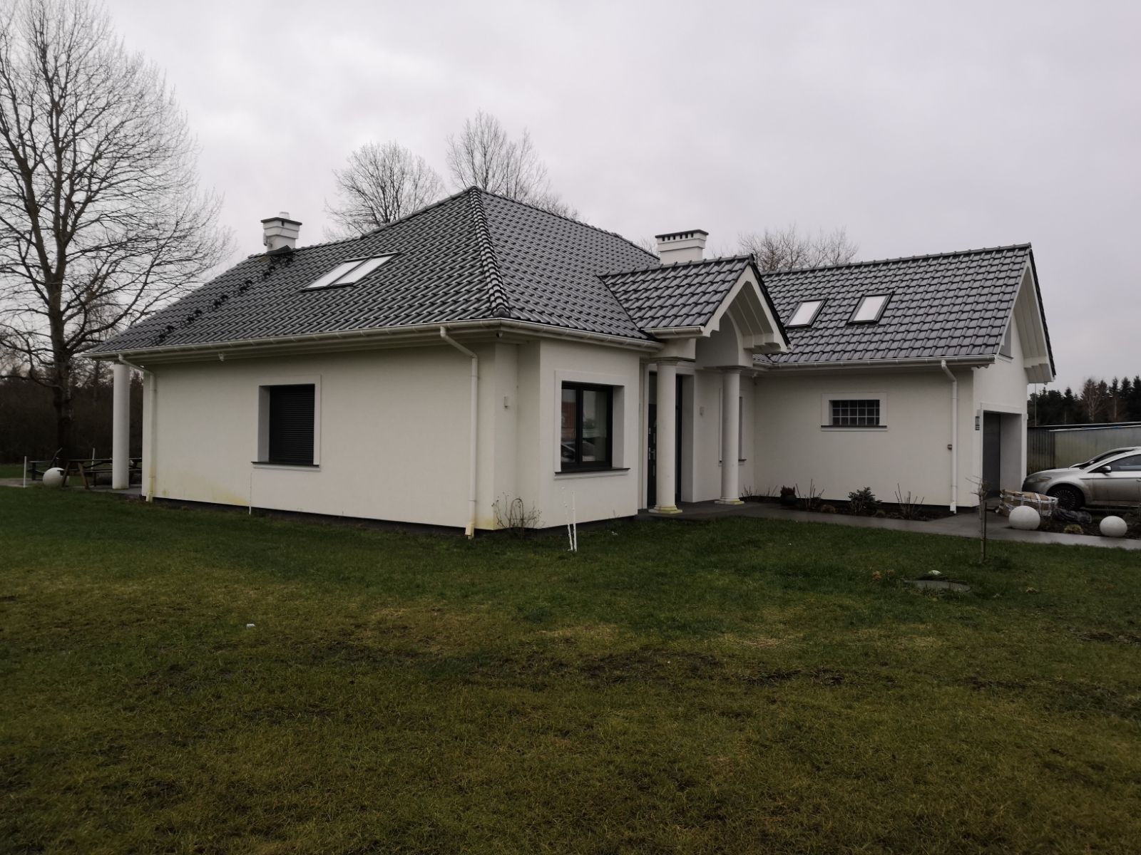 Nowoczesny, parterowy dom murowany z grafitową dachówką i białymi ścianami, widoczny z perspektywy trawnika w pochmurny dzień.
