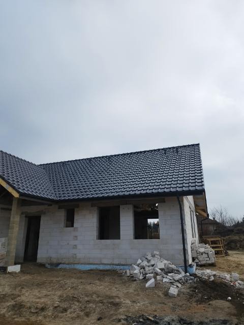 Surowy stan domu z białego pustaka, czarny dach z dachówki, stos pustaków przed ścianą, w tle pochmurne niebo.