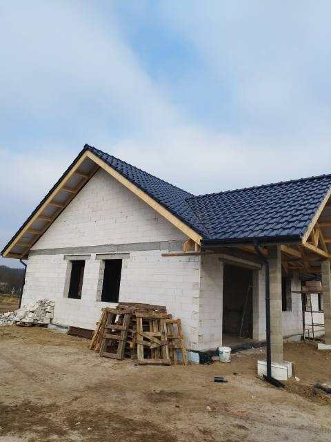 Niedokończony budynek z białych bloczków z ciemną dachówką, widoczne drewniane elementy konstrukcyjne dachu, okna bez ram, palety i materiały budowlane na placu budowy.