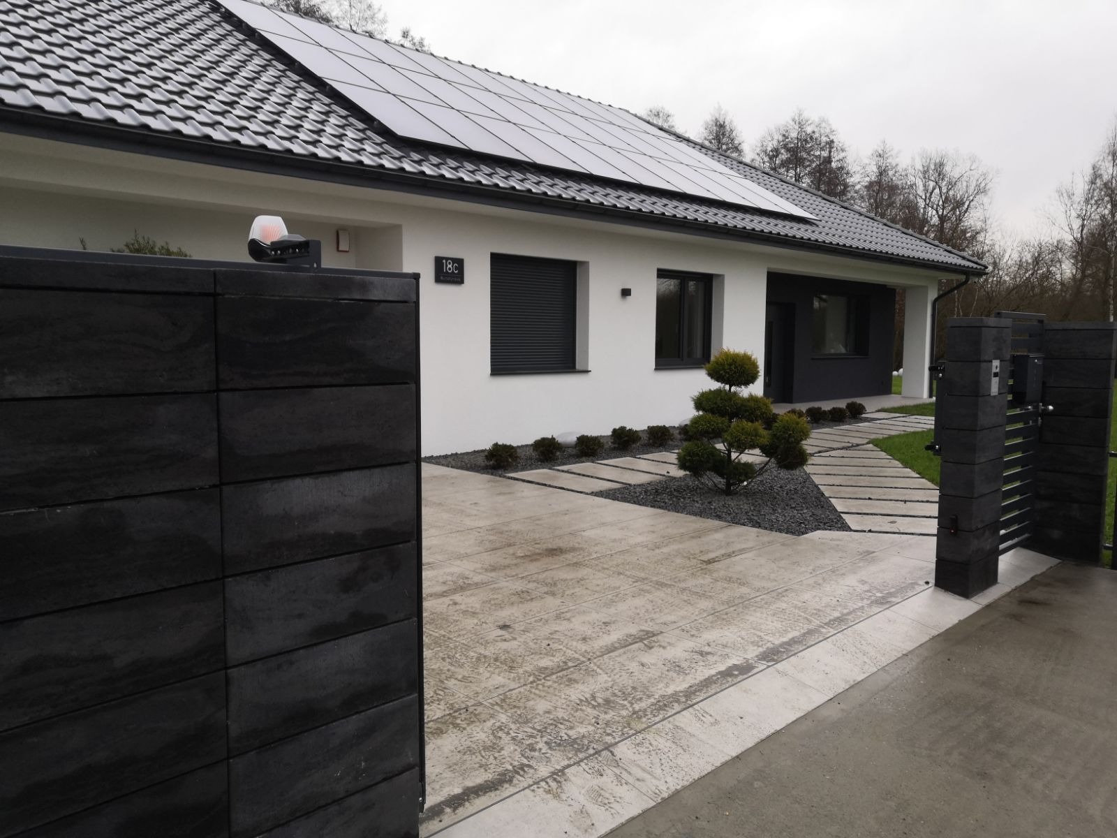 Nowoczesny dom jednorodzinny z panelami słonecznymi na dachu, czarnym ogrodzeniem z przesuwną bramą, podjazdem z betonowych płyt i ozdobnym krzewem.