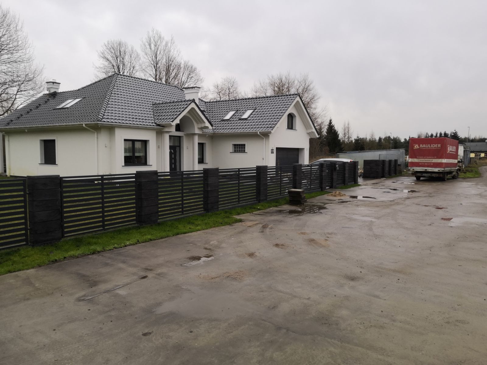 Nowoczesny dom murowany z szarą dachówką i ciemnym ogrodzeniem panelowym, widoczny w pochmurny dzień, z ciężarówką firmy budowlanej w tle.