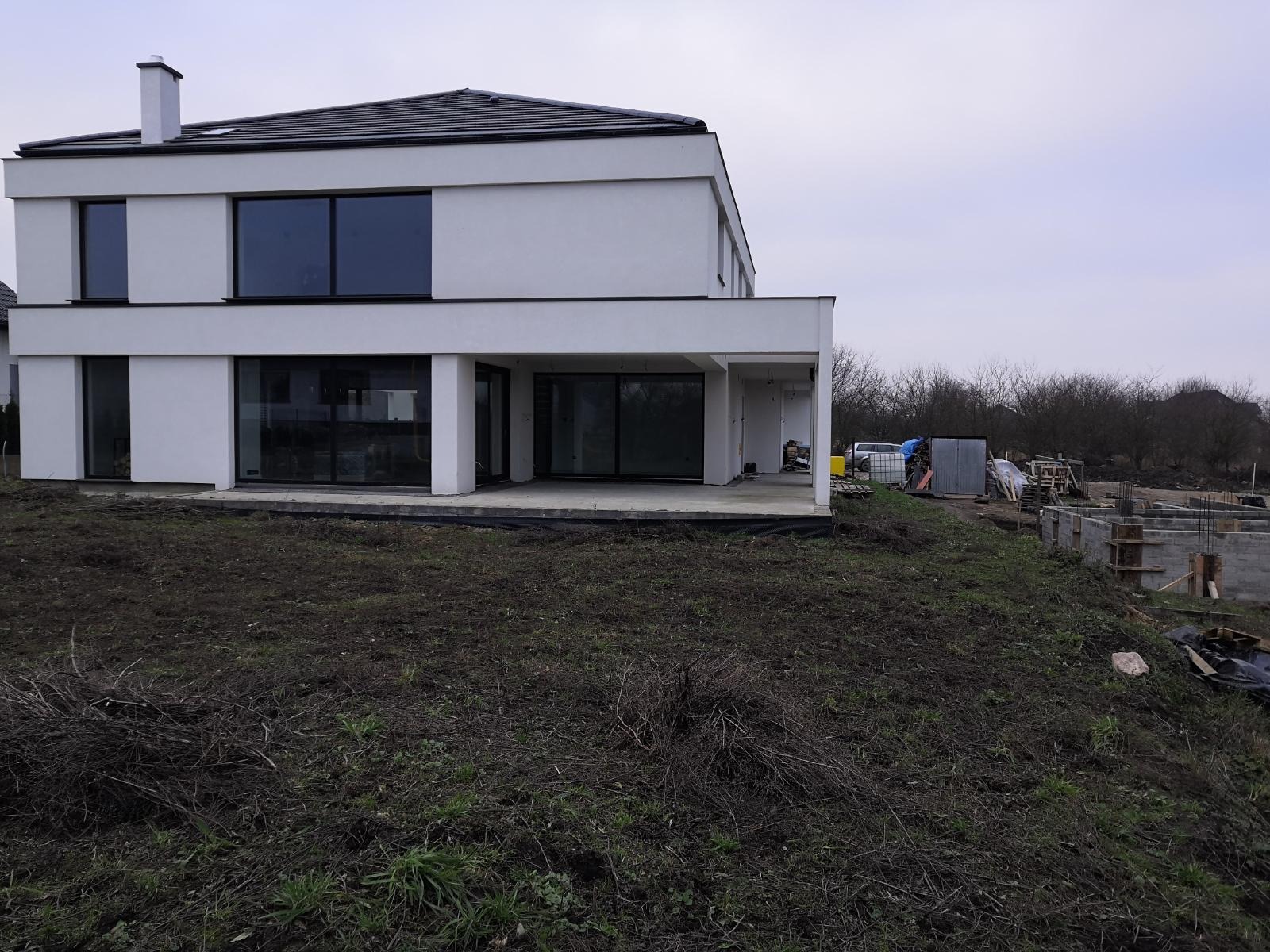 Dwukondygnacyjny, biały dom z płaskim dachem w trakcie budowy; widoczne fundamenty i teren budowy z materiałami.