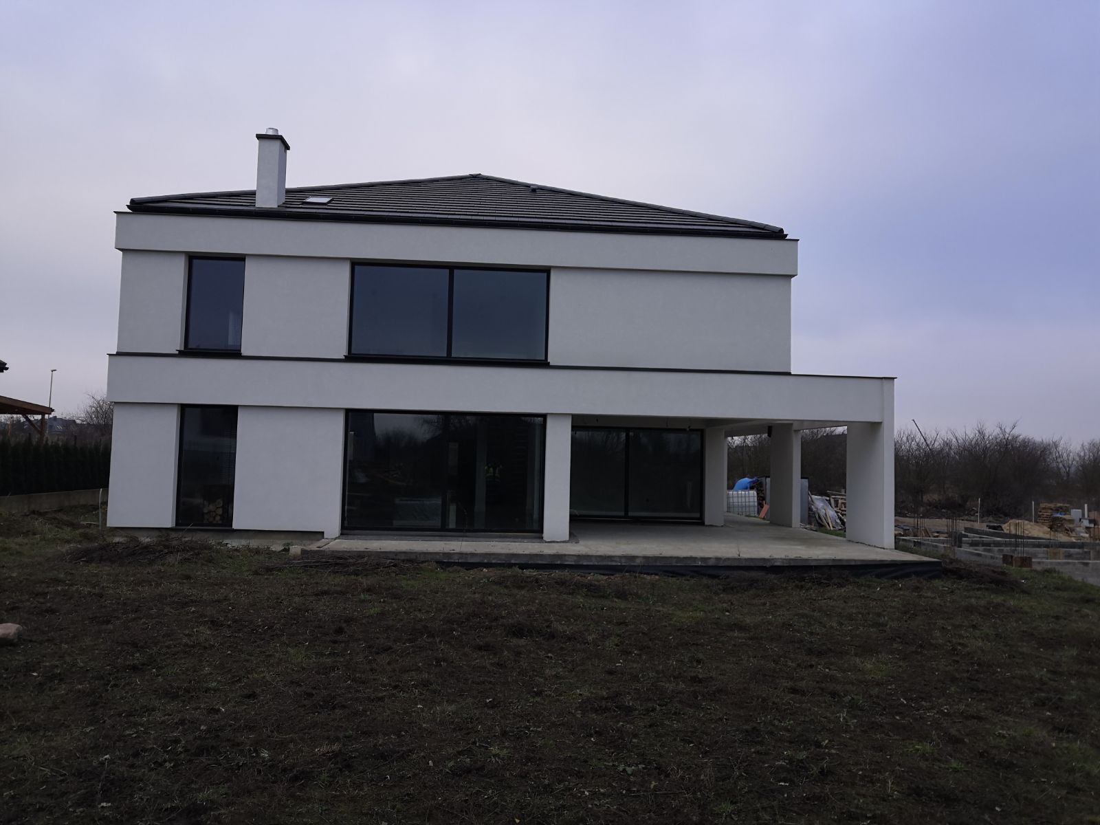 Dwukondygnacyjny dom murowany z płaskim dachem i dużymi oknami, w trakcie wykańczania elewacji zewnętrznej, widoczne fundamenty pod taras.