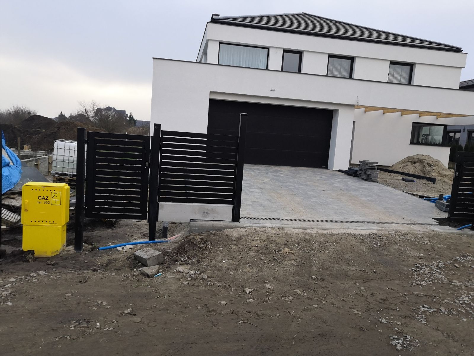 Nowoczesny dom murowany z czarną bramą garażową i poziomym ogrodzeniem, kostka brukowa na podjeździe, skrzynka gazowa przy ogrodzeniu.