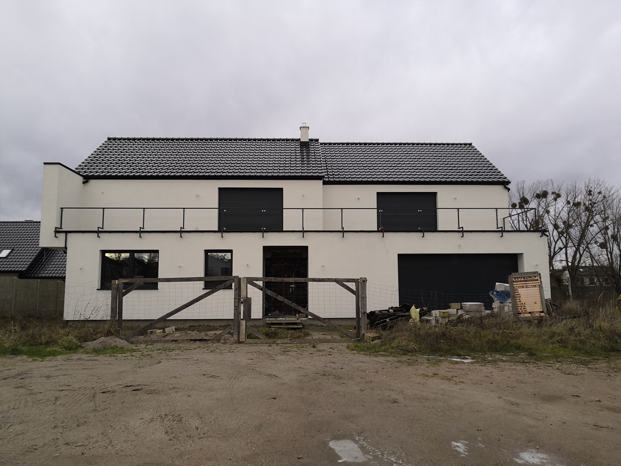 Nowoczesny dom z białymi ścianami i czarnym dachem z dachówki, widziany z zewnątrz, z drewnianym ogrodzeniem w pierwszym planie i materiałami budowlanymi obok garażu.