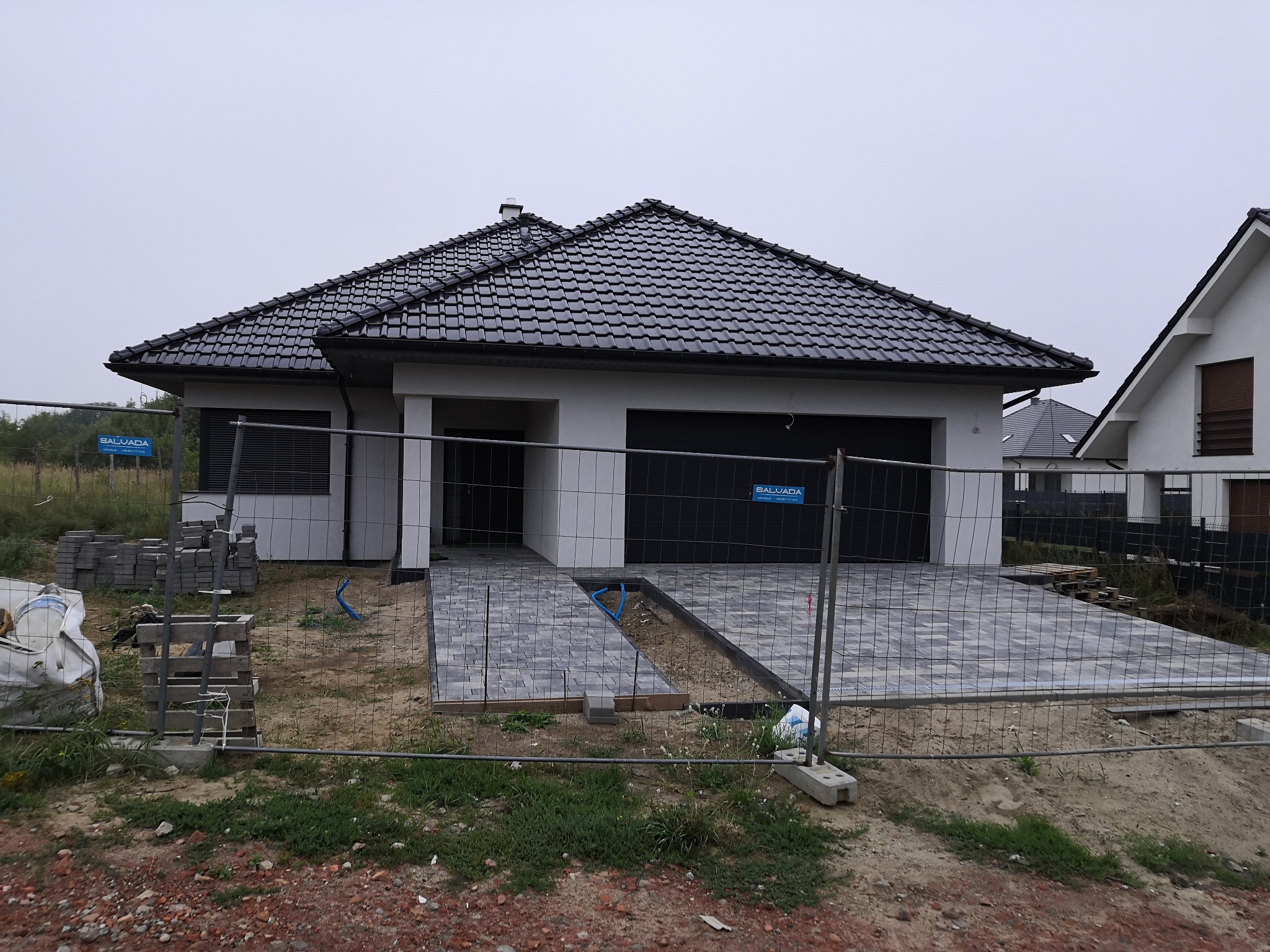 Parterowy dom jednorodzinny z czarną dachówką i wybrukowanym podjazdem z szarej kostki, w trakcie prac wykończeniowych, ogrodzony tymczasowym płotem.