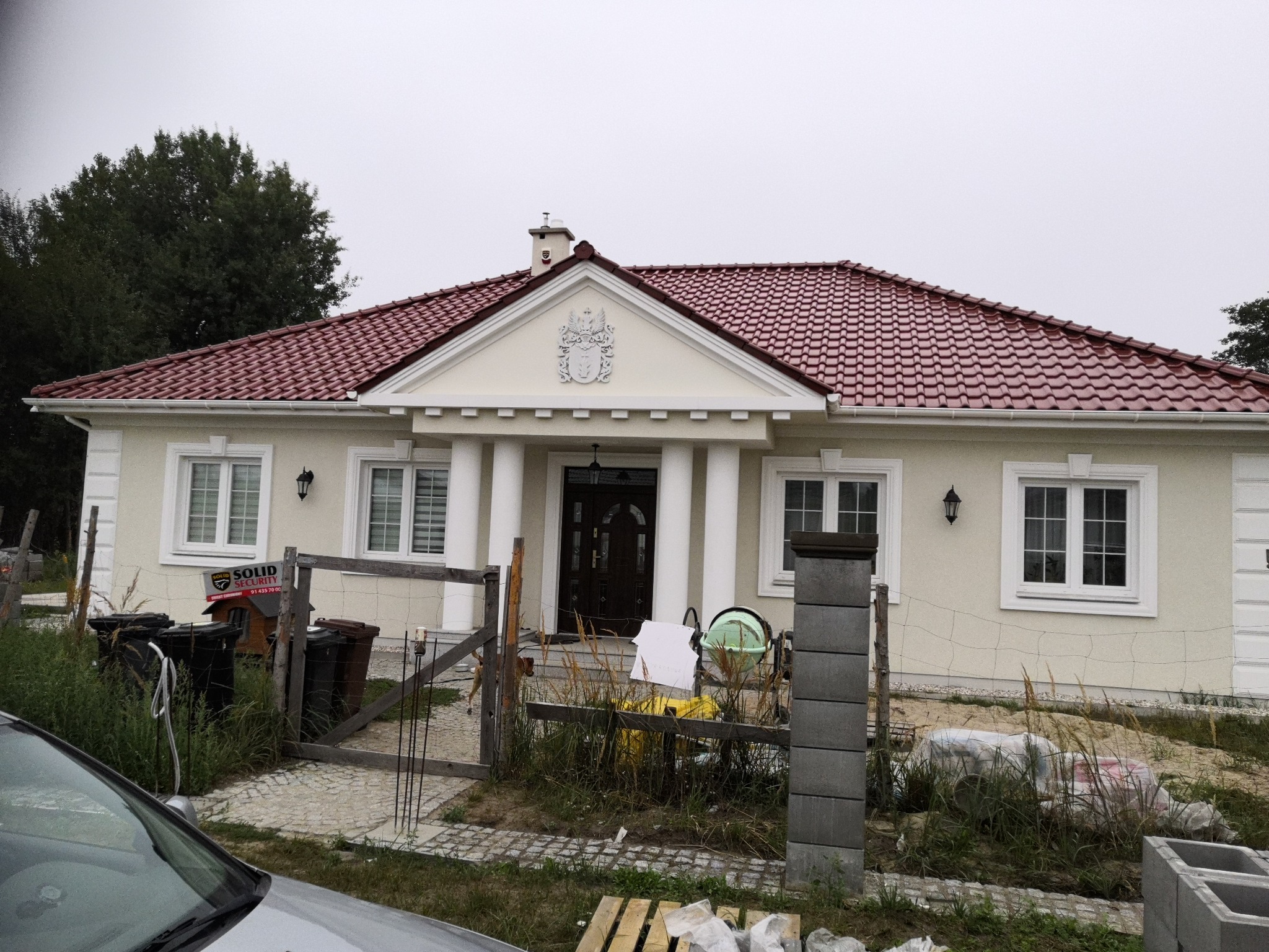 Parterowy dom z czerwoną dachówką i kolumnami przy wejściu, widoczne ślady budowy, ogrodzenie z siatki, gruz i materiały budowlane na posesji.