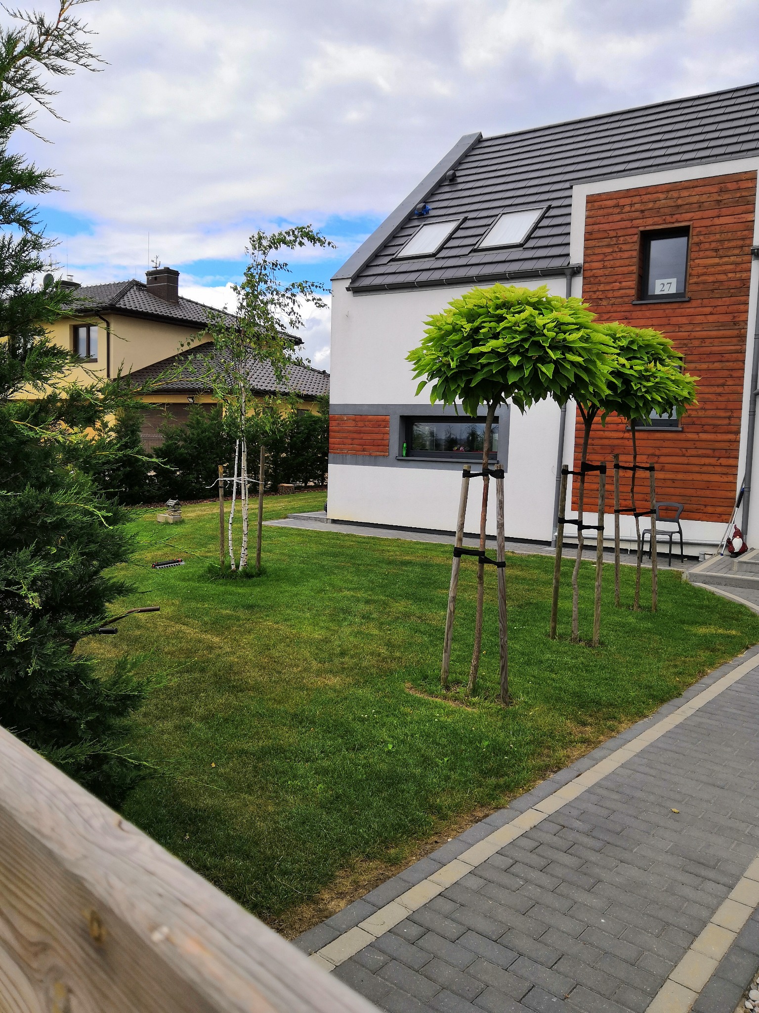 Nowoczesny dom z elementami drewnianymi, zadbany trawnik z młodymi drzewkami z podporami, kostka brukowa, widok z zewnątrz.