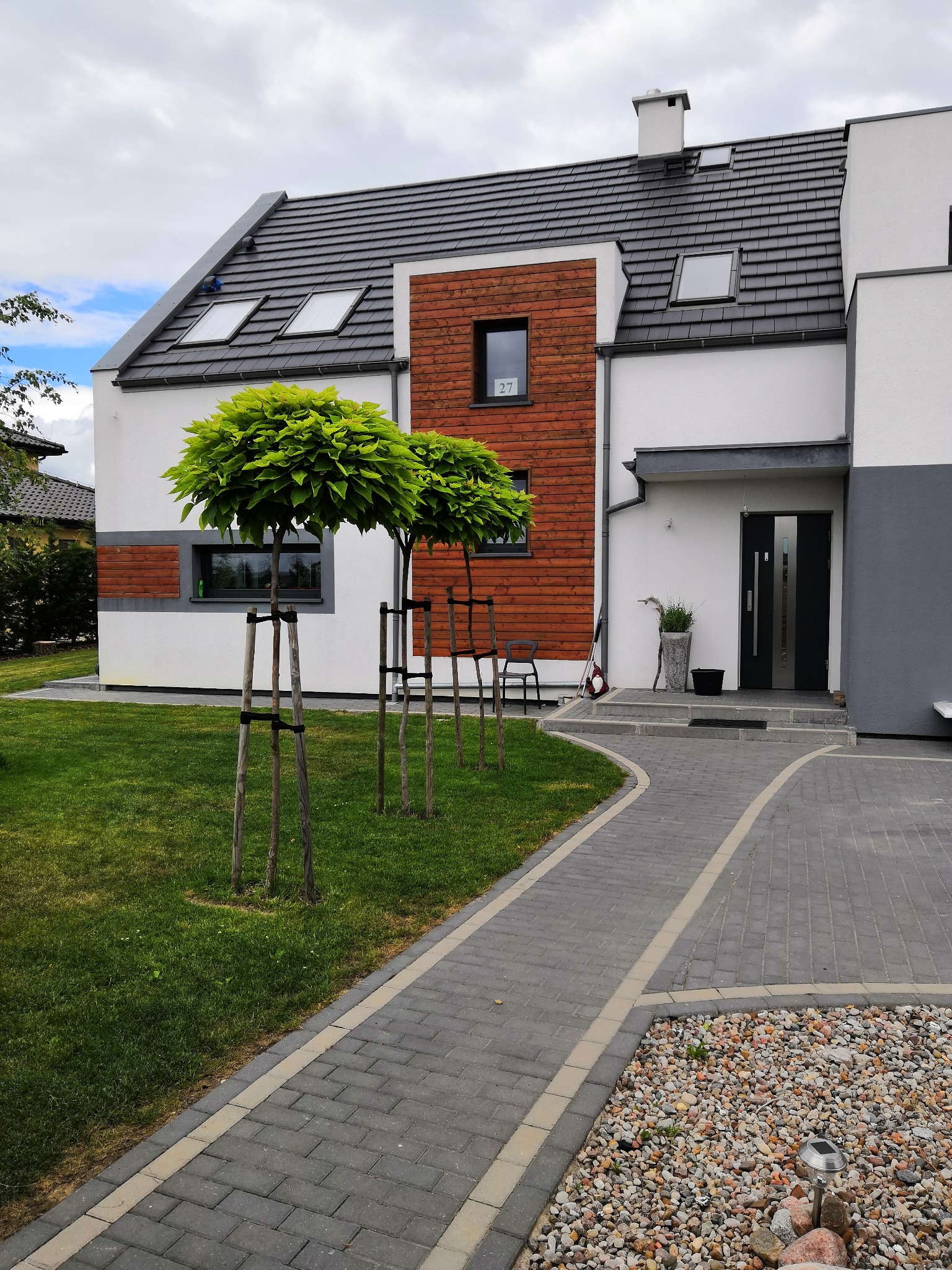 Nowoczesny dom jednorodzinny z drewnianą elewacją, szarą dachówką i oknami dachowymi, otoczony trawnikiem i ozdobnymi drzewkami na pniu, z brukowanym chodnikiem prowadzącym do wejścia.