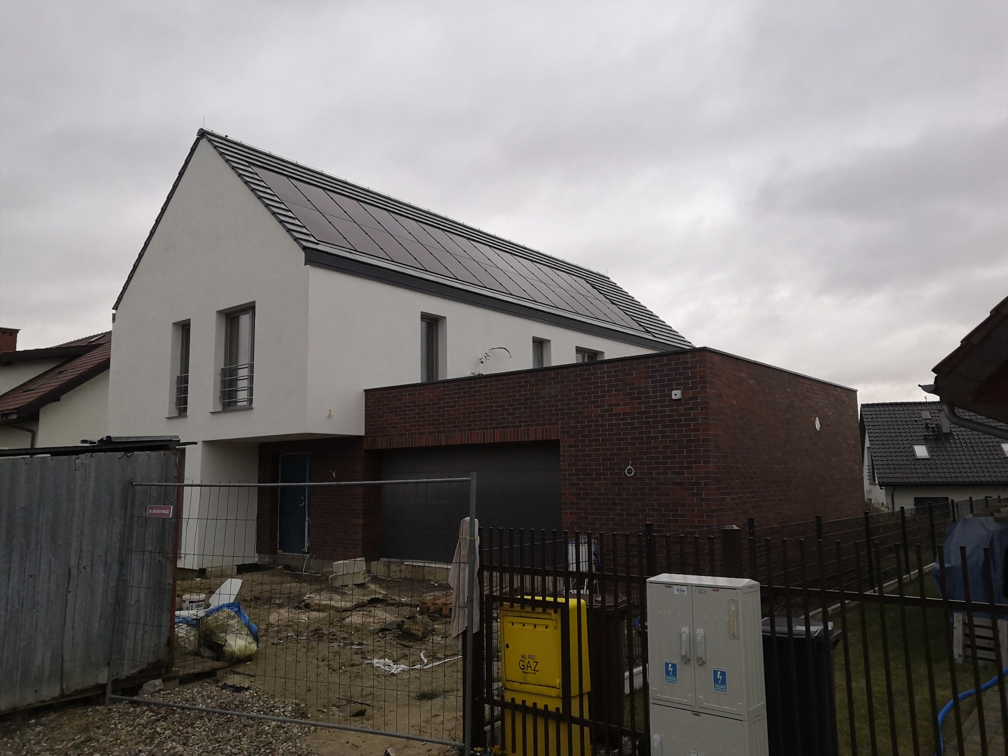Nowoczesny dom jednorodzinny z panelami słonecznymi na dachu, częściowo wykończony, z widocznym ogrodzeniem budowlanym i skrzynką gazową przed posesją, sfotografowany w pochmurny dzień.