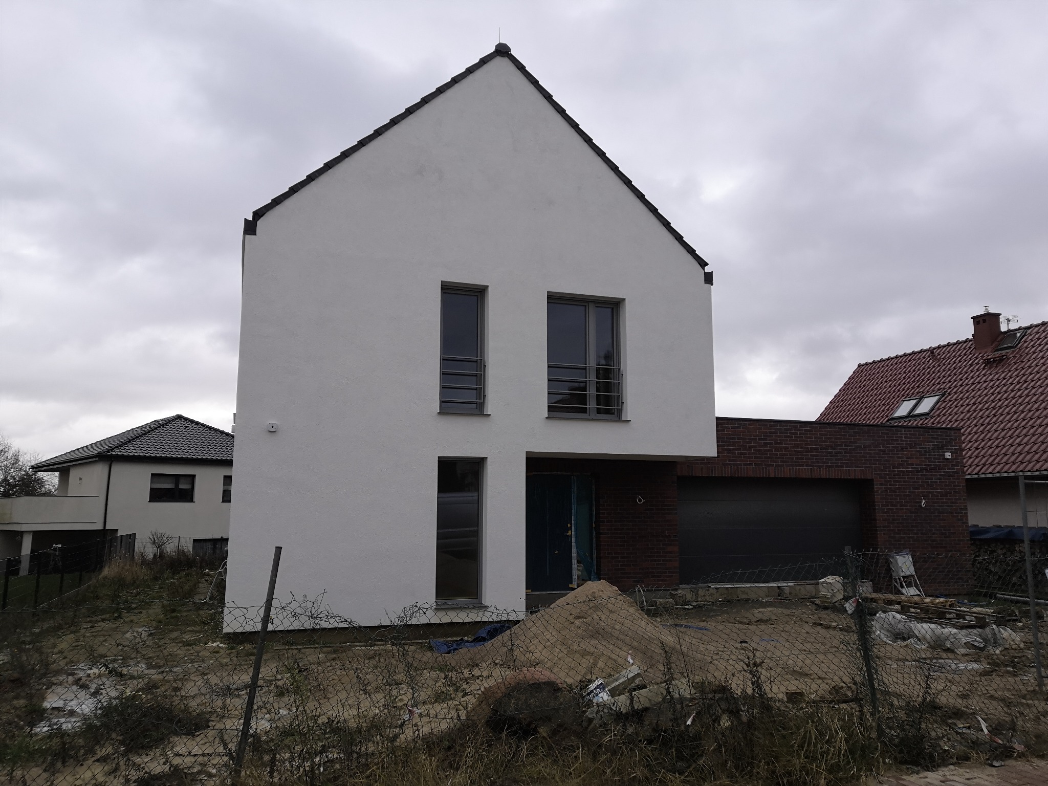 Nowoczesny dom z białymi ścianami i ciemną stolarką okienną, widoczny w stanie surowym zamkniętym, z widocznym stosem piasku przed wejściem i niezagospodarowanym terenem wokół.