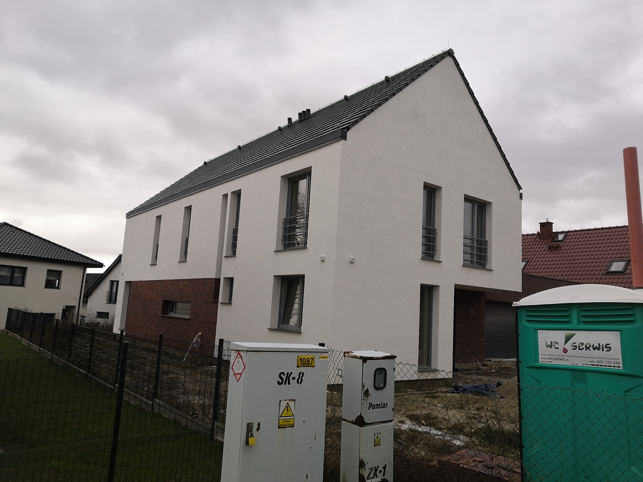 Nowoczesny, dwukondygnacyjny dom z białym tynkiem i elementami cegły klinkierowej, czarny dach, widok z zewnątrz, na pierwszym planie skrzynki elektryczne i toaleta przenośna.