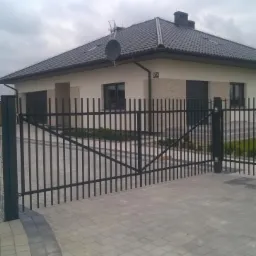 Nowoczesny dom z ciemną dachówką i kremową elewacją otoczony czarnym, metalowym ogrodzeniem panelowym i bramą wjazdową, widok z zewnątrz posesji.