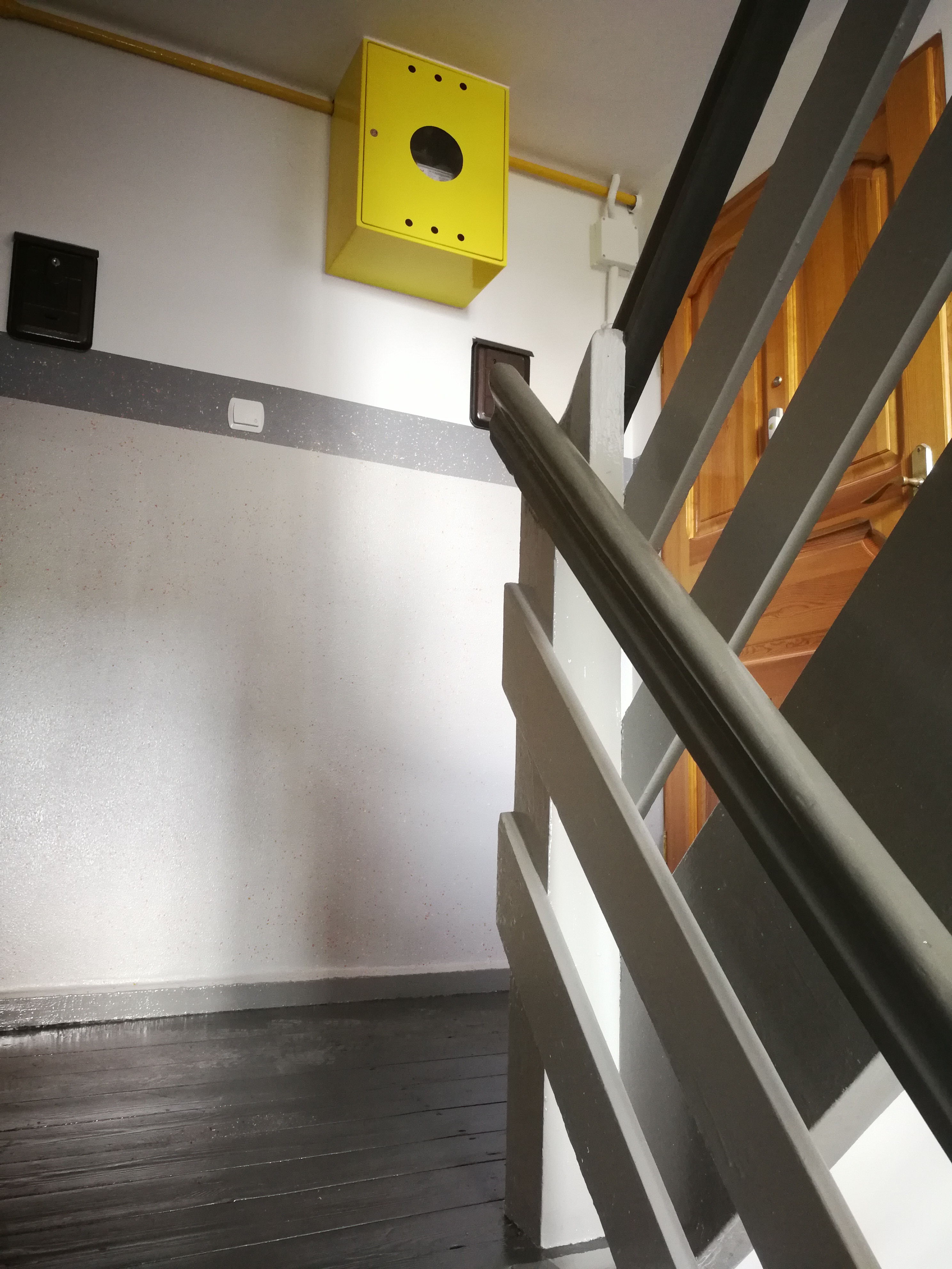 Klatka schodowa po odświeżeniu: pomalowane ściany na biało z szarym pasem, ciemne drewniane schody i szare barierki, żółta skrzynka na ścianie.