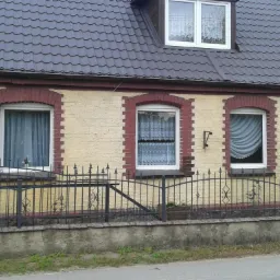 Elewacja domu z żółtą fasadą i ceglanymi obramowaniami okien. Widoczne okna z firankami, metalowe ogrodzenie i numer domu.