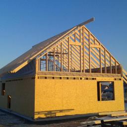 Arkadiusz Smogulecki - Budowa domu w stanie surowym otwartym: ściany z płyty OSB, drewniana konstrukcja dachu na tle błękitnego nieba i sąsiednich domów. Słońce oświetla budowę.