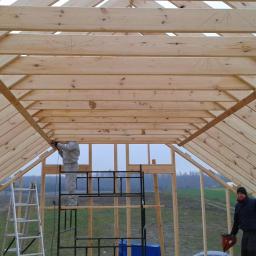 Arkadiusz Smogulecki - Konstrukcja drewniana dachu w trakcie budowy, widok od wewnątrz. Pracownik montuje elementy na rusztowaniu, drugi stoi na ziemi z narzędziem.