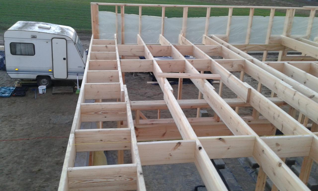 Drewniany szkielet konstrukcji domu w budowie, widok z góry. Na zewnątrz przyczepa kempingowa i narzędzia. Surowy stan, detale łączeń belek.