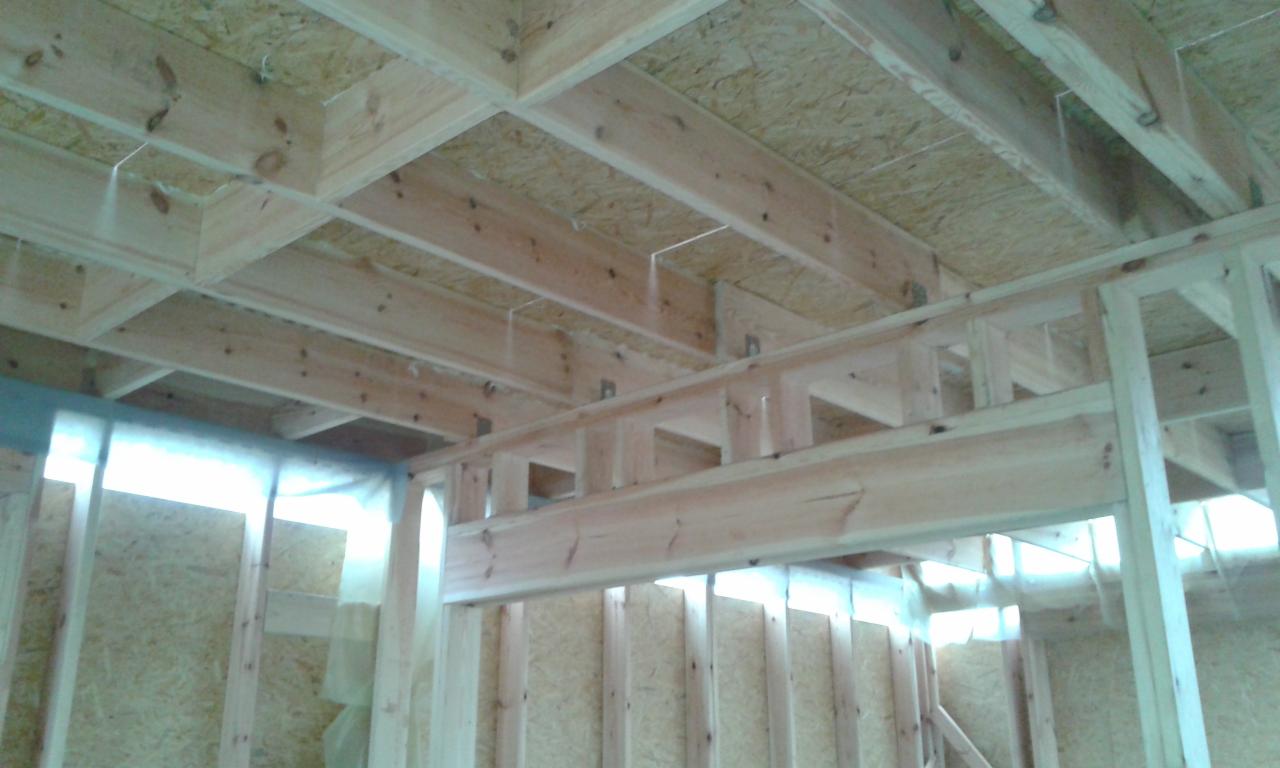 Drewniana konstrukcja szkieletowa budynku w trakcie budowy, widoczne belki stropowe i ściany z płyty OSB, zabezpieczone folią. Surowy stan, jasne drewno.