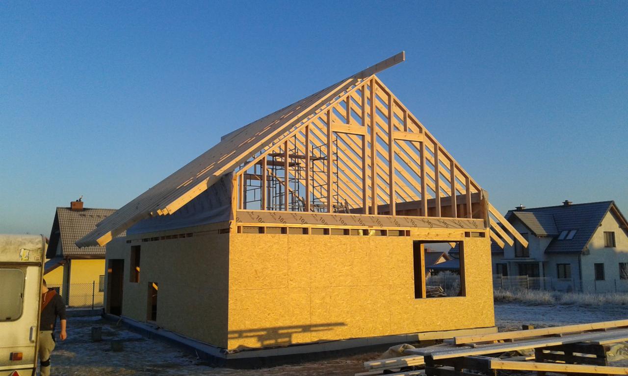Budowa domu w stanie surowym otwartym: ściany z płyty OSB, drewniana konstrukcja dachu na tle błękitnego nieba i sąsiednich domów. Słońce oświetla budowę.