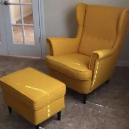 Musztardowy fotel uszak z podnóżkiem, ustawiony na szarych płytkach podłogowych, widoczne drzwi balkonowe w tle