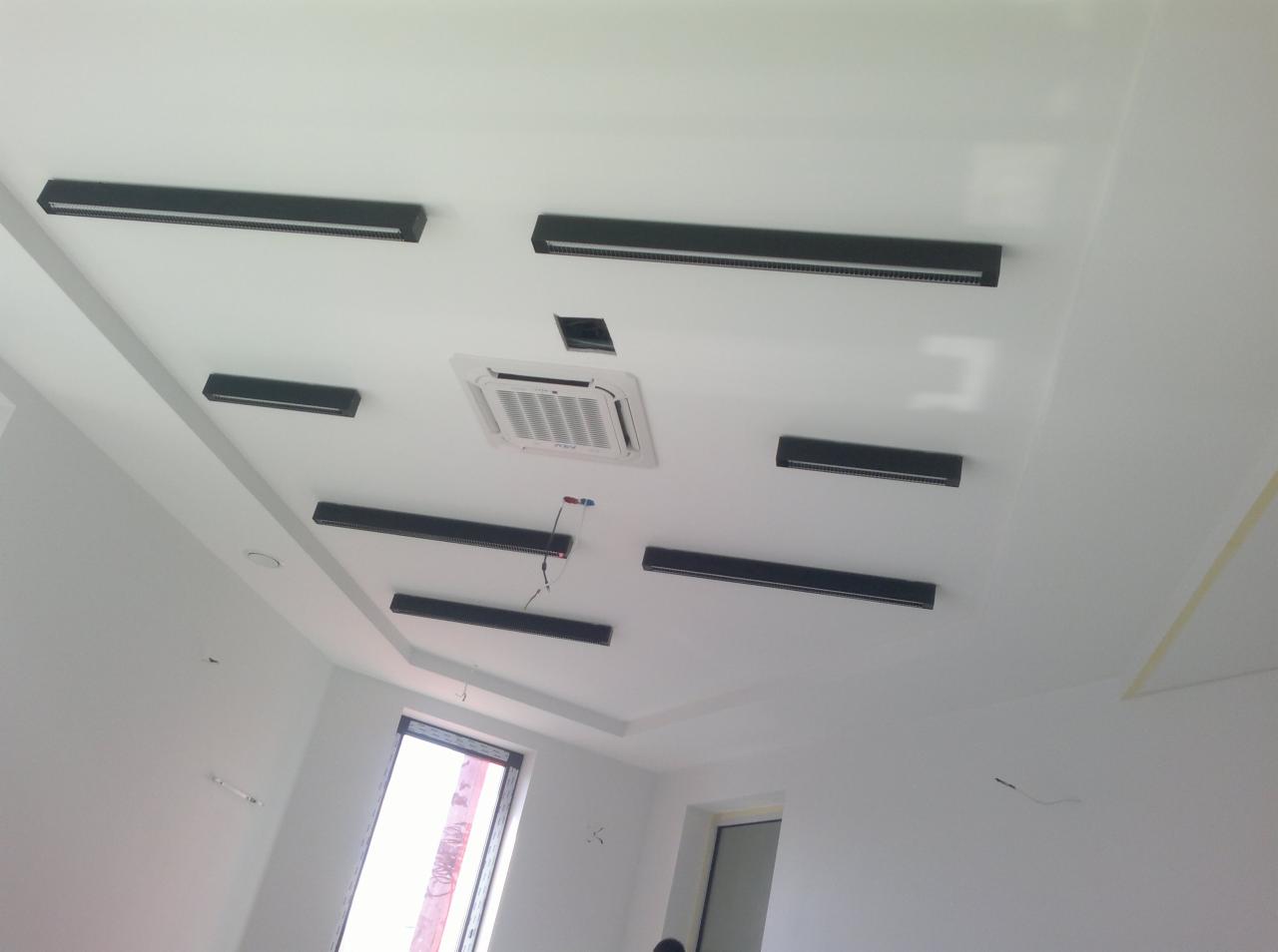 Biały sufit z zamontowanymi czarnymi, podłużnymi lampami LED i klimatyzacją kasetonową. Widok z dołu, kątowe ujęcie, widoczne okna i fragmenty ścian.