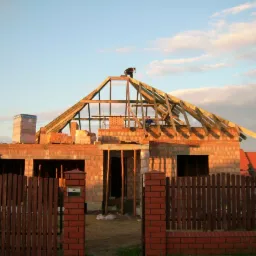 Budowa domu jednorodzinnego z czerwonej cegły: wzniesione ściany, więźba dachowa w trakcie montażu, murarz na dachu. Widok zza drewnianego ogrodzenia.