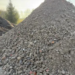 Kupa szarego, pokruszonego materiału budowlanego z fragmentami czerwonej cegły, uformowana w stożek na tle zieleni i błękitnego nieba z lekkim zachmurzeniem.