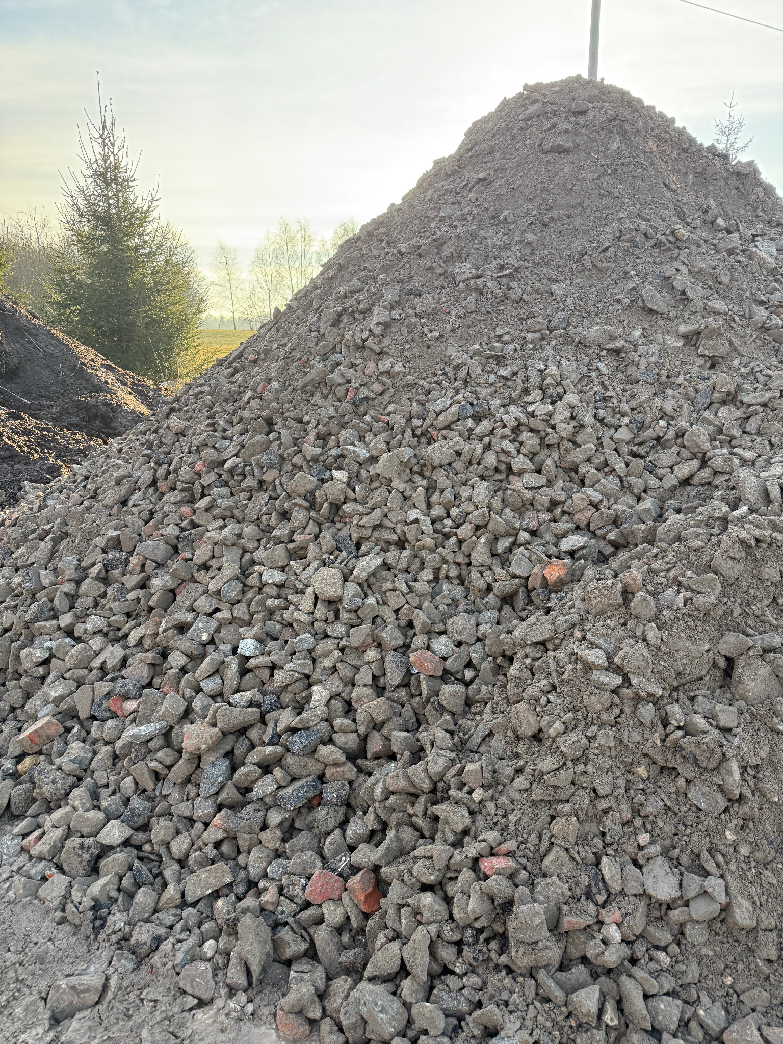 Kupa szarego, pokruszonego materiału budowlanego z fragmentami czerwonej cegły, uformowana w stożek na tle zieleni i błękitnego nieba z lekkim zachmurzeniem.