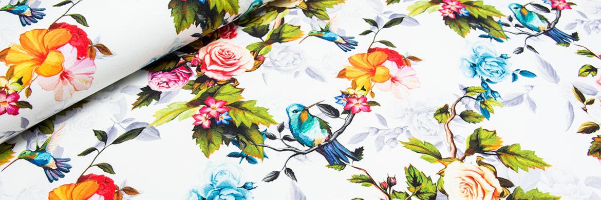 Zwinięty fragment tkaniny z nadrukiem w róże, kolibry i inne kwiaty na białym tle, z widoczną fakturą materiału.