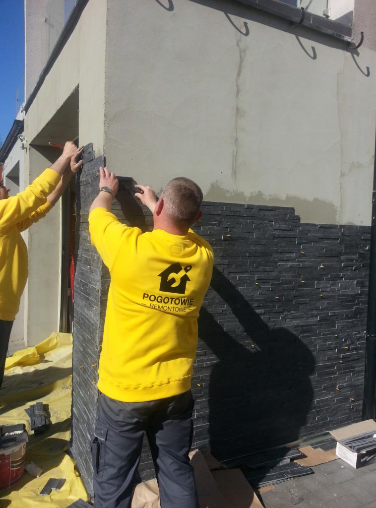 Prace wykończeniowe elewacji: dwóch mężczyzn w żółtych koszulkach układa czarne płytki imitujące kamień na ścianie budynku. Widoczne narzędzia i materiały budowlane.