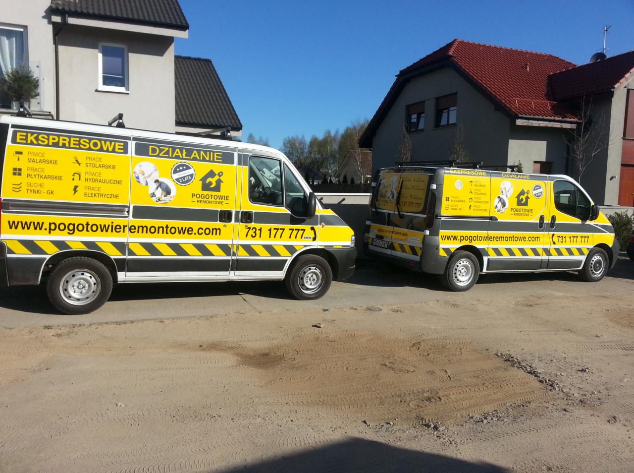 Dwa busy 'Pogotowie Remontowe' z żółto-szarym oklejeniem, zaparkowane na żwirowym placu przed domami. Na busach widoczne usługi: malarskie, hydrauliczne, elektryczne.