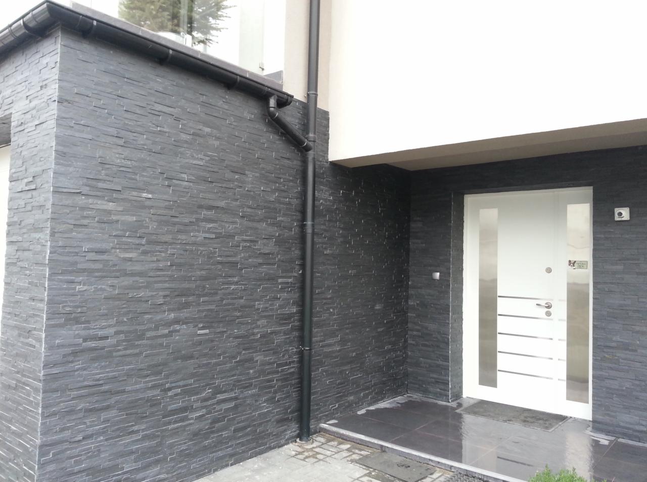 Elewacja z ciemnego kamienia łupanego, z czarną rynną i białą, nowoczesną stolarką drzwiową. Widoczny fragment chodnika z kostki brukowej.