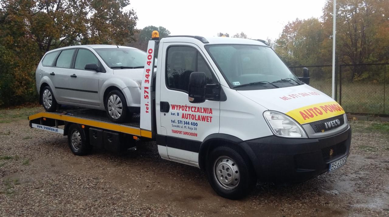 Biała laweta Iveco z załadowanym srebrnym kombi Ford Focus, widoczne logo firmy 'Wypożyczalnia Autolawet' i dane kontaktowe na boku pojazdu.