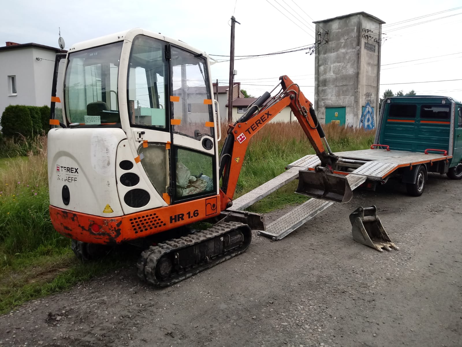 Mała koparka Terex HR 1.6 na lawecie transportowej, obok dodatkowa łyżka, w tle budynek transformatora i zabudowa jednorodzinna.