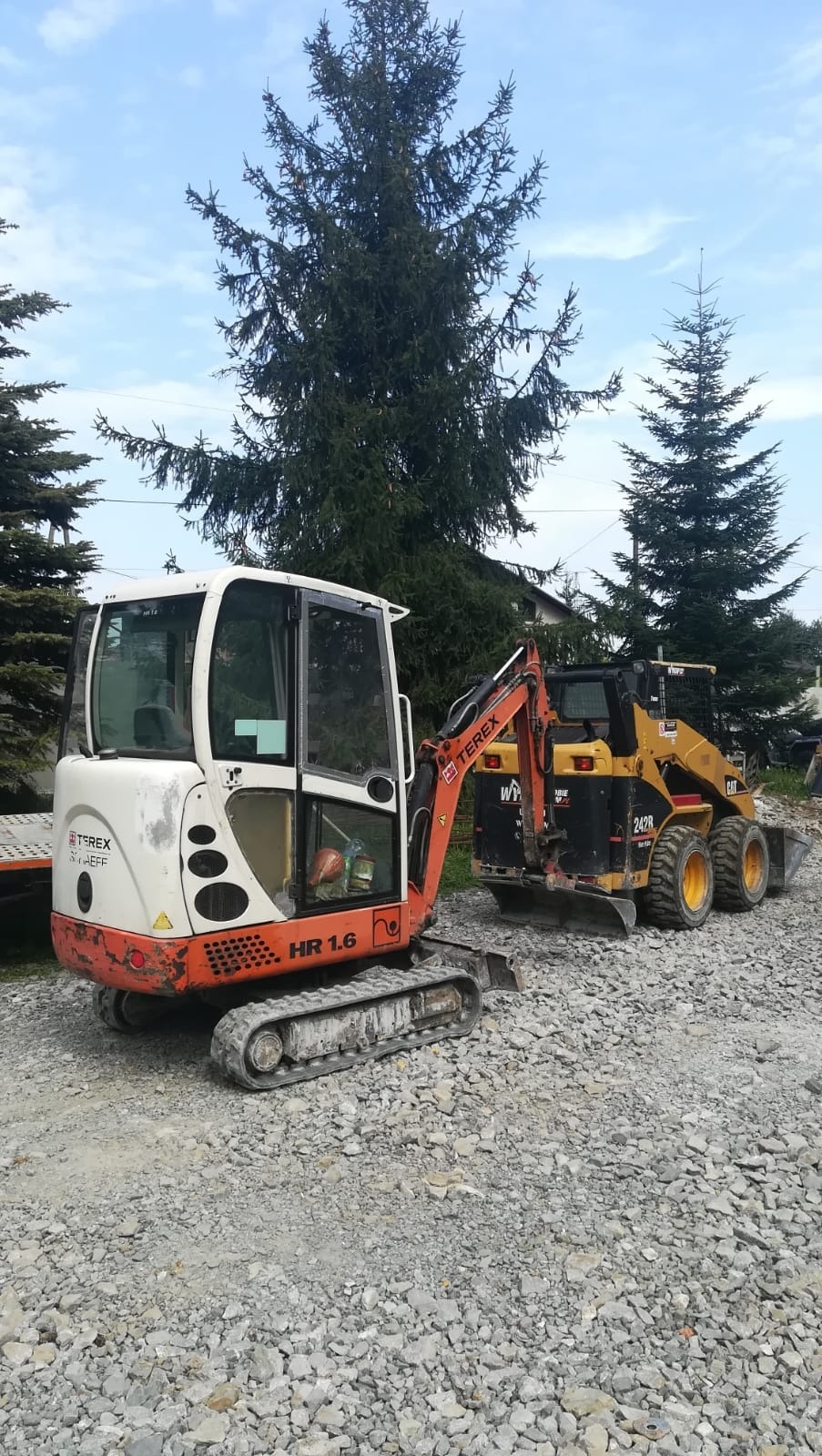 Mini koparka Terex HR 1.6 i ładowarka Caterpillar 242R na żwirowym podłożu, w tle iglaste drzewa i błękitne niebo.