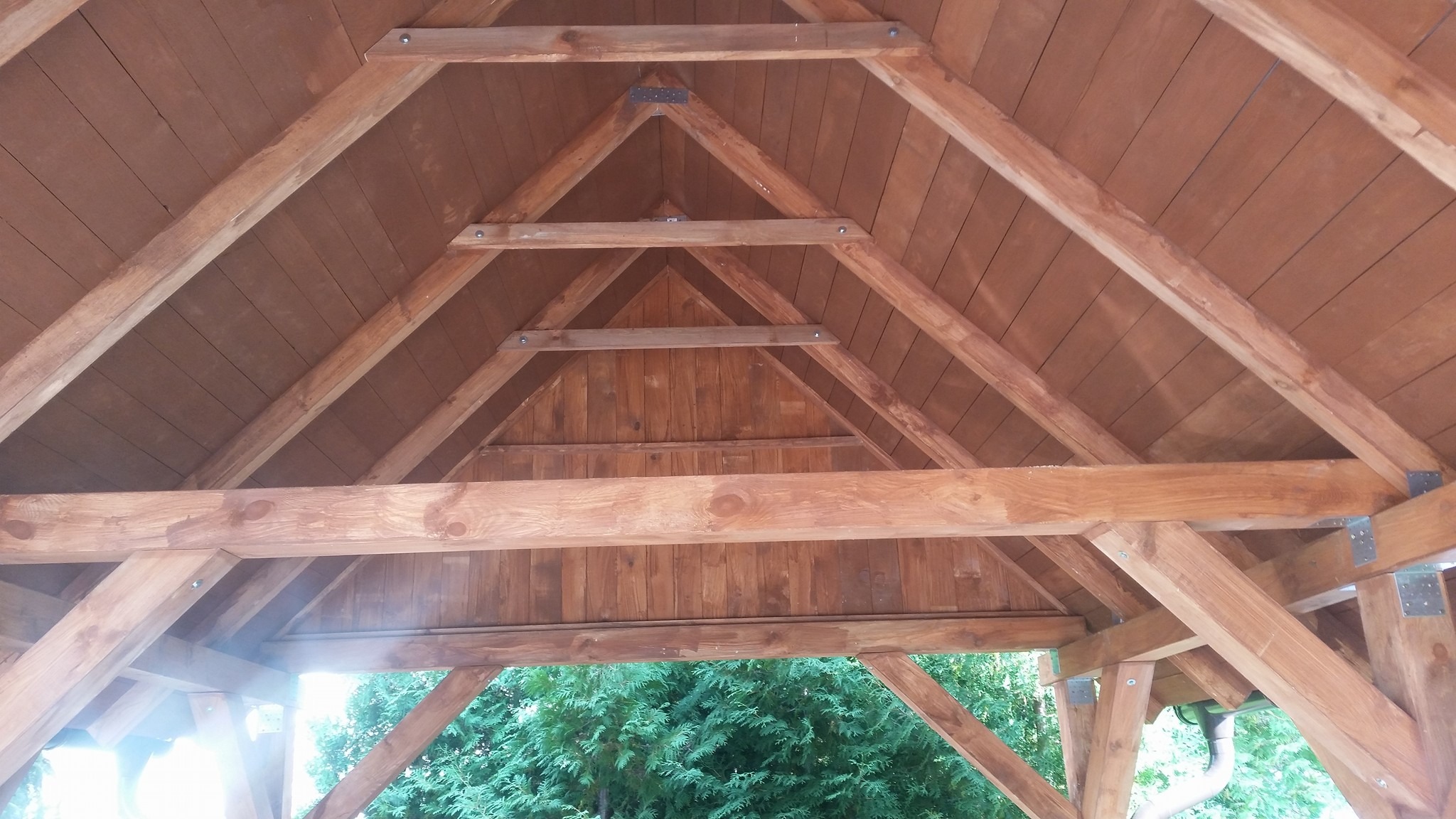 Wnętrze drewnianej konstrukcji z widocznym dachem i elementami nośnymi, połączonymi metalowymi płytkami, z zielenią w tle.