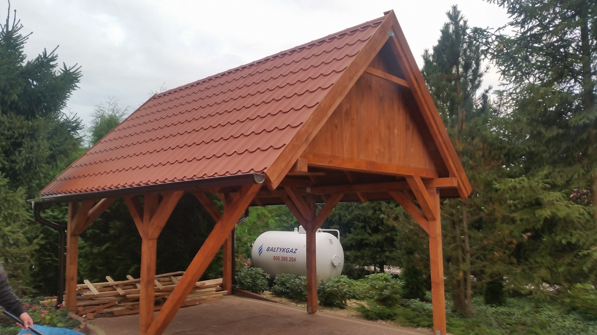 Zadaszona wiata z drewnianą konstrukcją i czerwoną dachówką, w tle zbiornik na gaz i zielona roślinność.
