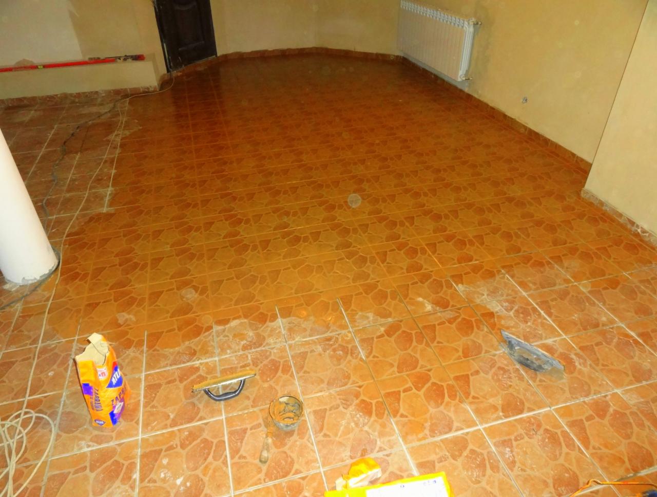 Proces fugowania brązowych, nieregularnych płytek podłogowych w pokoju. Widoczny worek z fugą, narzędzia glazurnicze i częściowo zafugowana powierzchnia.