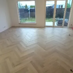 Jasny parkiet ułożony we wzór jodełki francuskiej w pustym pokoju z białymi ścianami i dużymi oknami balkonowymi, widoczne przygotowane materiały do wykończenia.