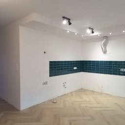 Agnieszka Szymańska Kujawa Pośrednictwo nieruchomości - Wnętrze w trakcie remontu: ułożona podłoga w jodełkę, fragment ściany z zielonymi płytkami i gniazdkami elektrycznymi, widoczne przewody i rury wentylacyjne pod sufitem.