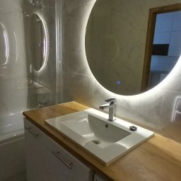 Agnieszka Szymańska Kujawa Pośrednictwo nieruchomości - Nowoczesna łazienka z okrągłym lustrem podświetlanym LED, białą umywalką wpuszczoną w drewniany blat i pralką Amica w zabudowie.