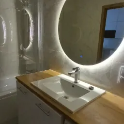 Nowoczesna łazienka z okrągłym lustrem podświetlanym LED, białą umywalką wpuszczoną w drewniany blat i pralką Amica w zabudowie.