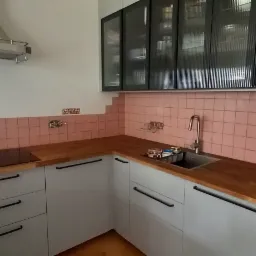 Fragment kuchni z różowymi płytkami ściennymi ułożonymi schodkowo, jasnoszare szafki z drewnianym blatem, czarne uchwyty, szklane szafki wiszące z czarnymi ramami, okap, piekarnik z naklejkami...