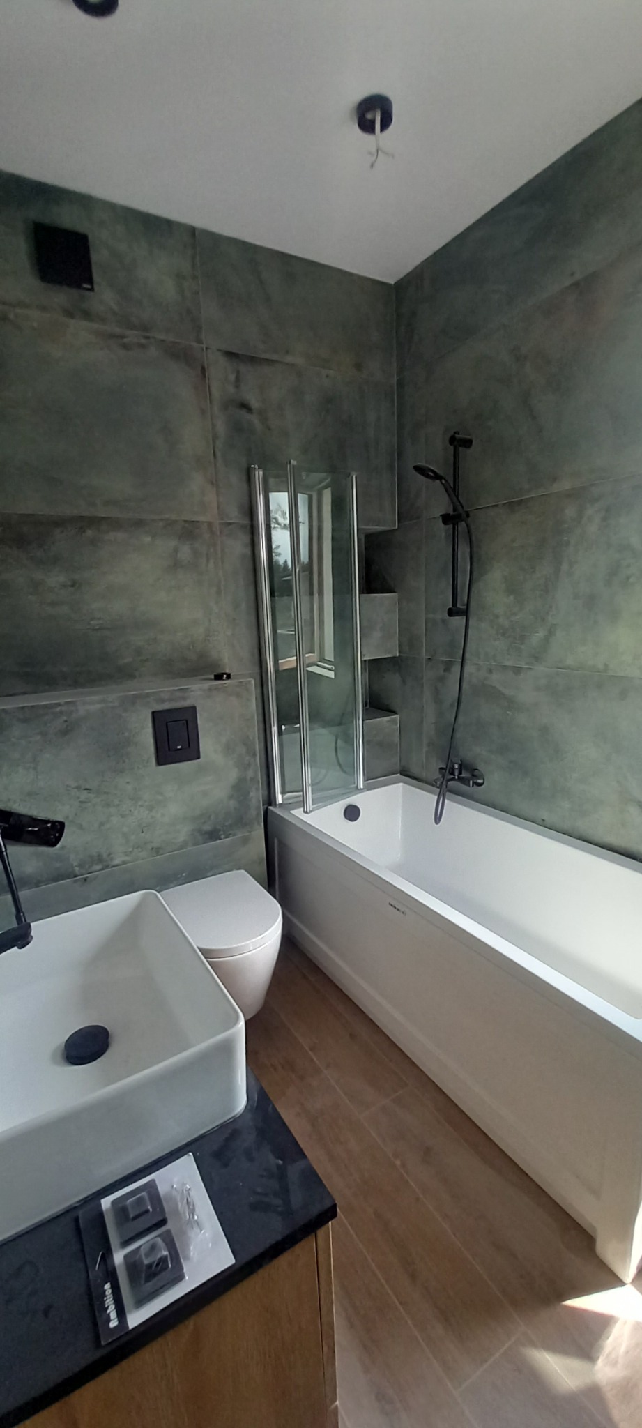 Nowoczesna łazienka z umywalką nablatową, białą wanną z parawanem nawannowym i płytkami imitującymi beton na ścianach. Widoczne niedokończone podłączenie oświetlenia sufitowego.