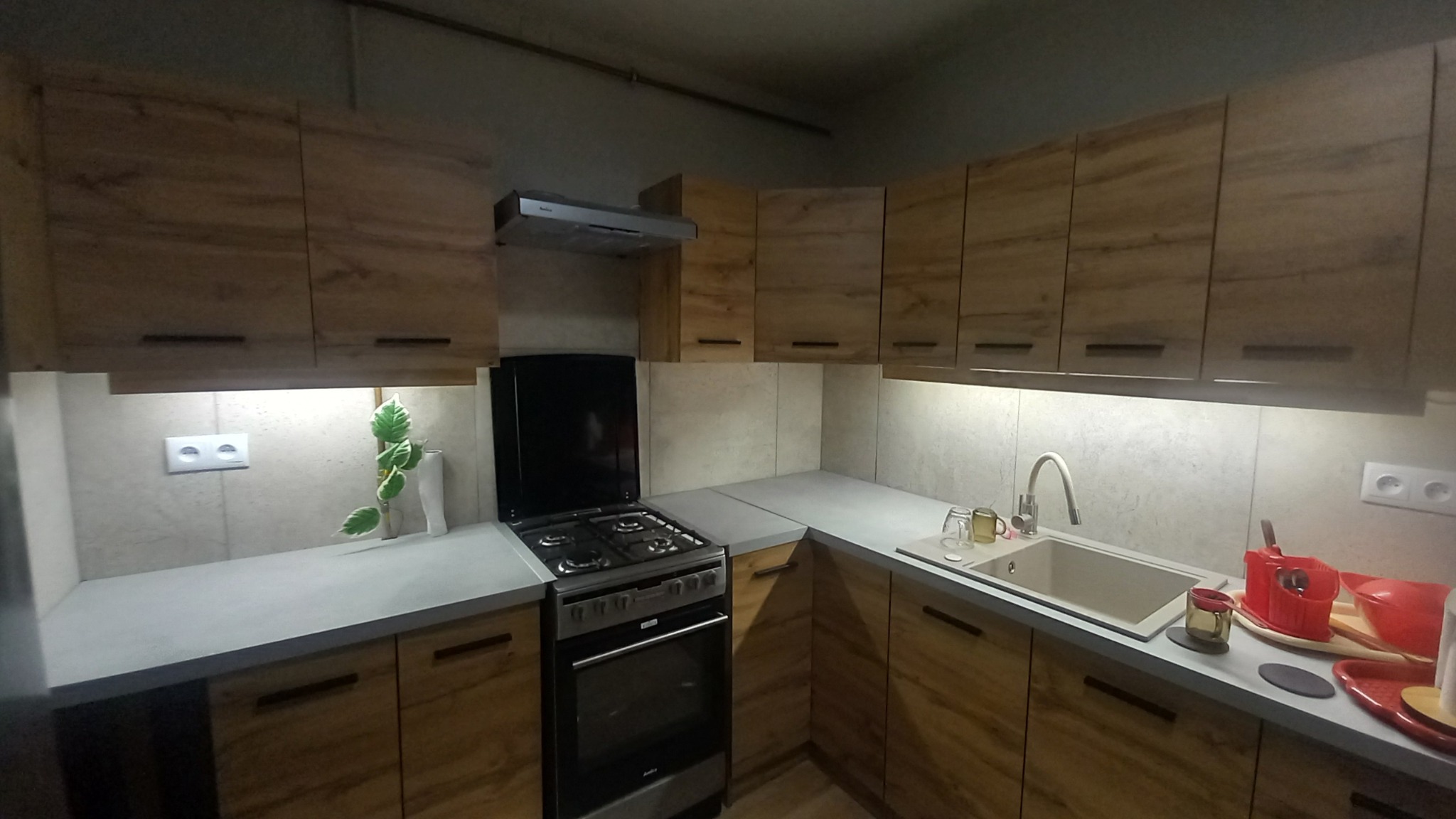 Nowoczesna kuchnia z drewnianymi szafkami, szarym blatem i płytkami w odcieniach beżu, oświetleniem podszafkowym i czarną kuchenką. Na blacie widoczne naczynia i dekoracyjna roślina.