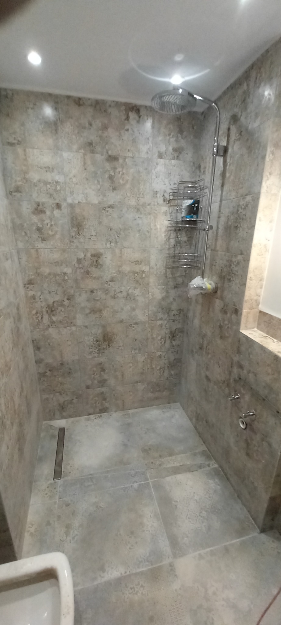 Ukończona kabina prysznicowa z szarymi, wzorzystymi płytkami ceramicznymi na ścianach i podłodze, wyposażona w deszczownicę, półkę na kosmetyki i odpływ liniowy.